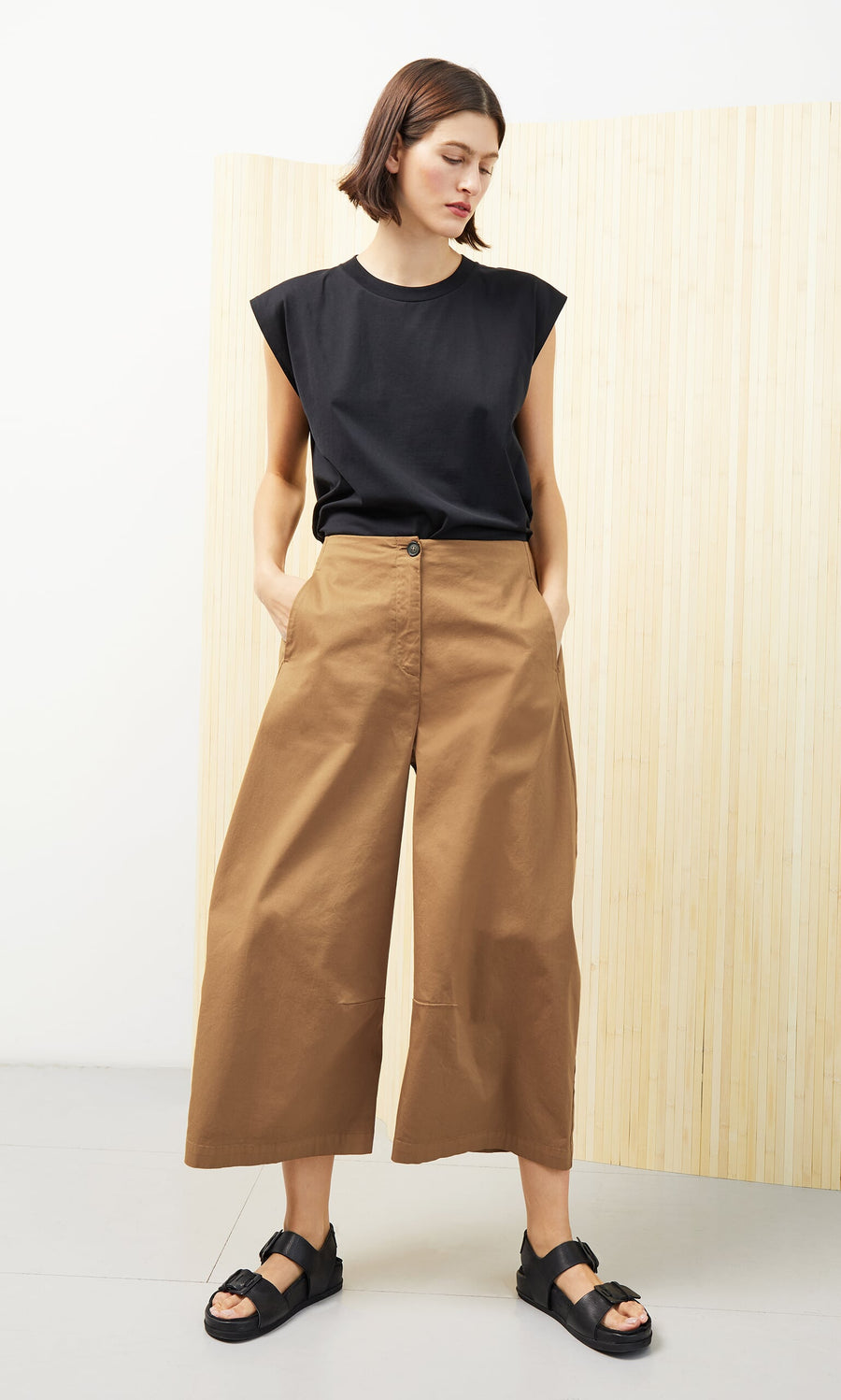 Plümo- Woman wearing Karate Pants, a black sleeveless top, and black sandals, standing with hands in pockets.