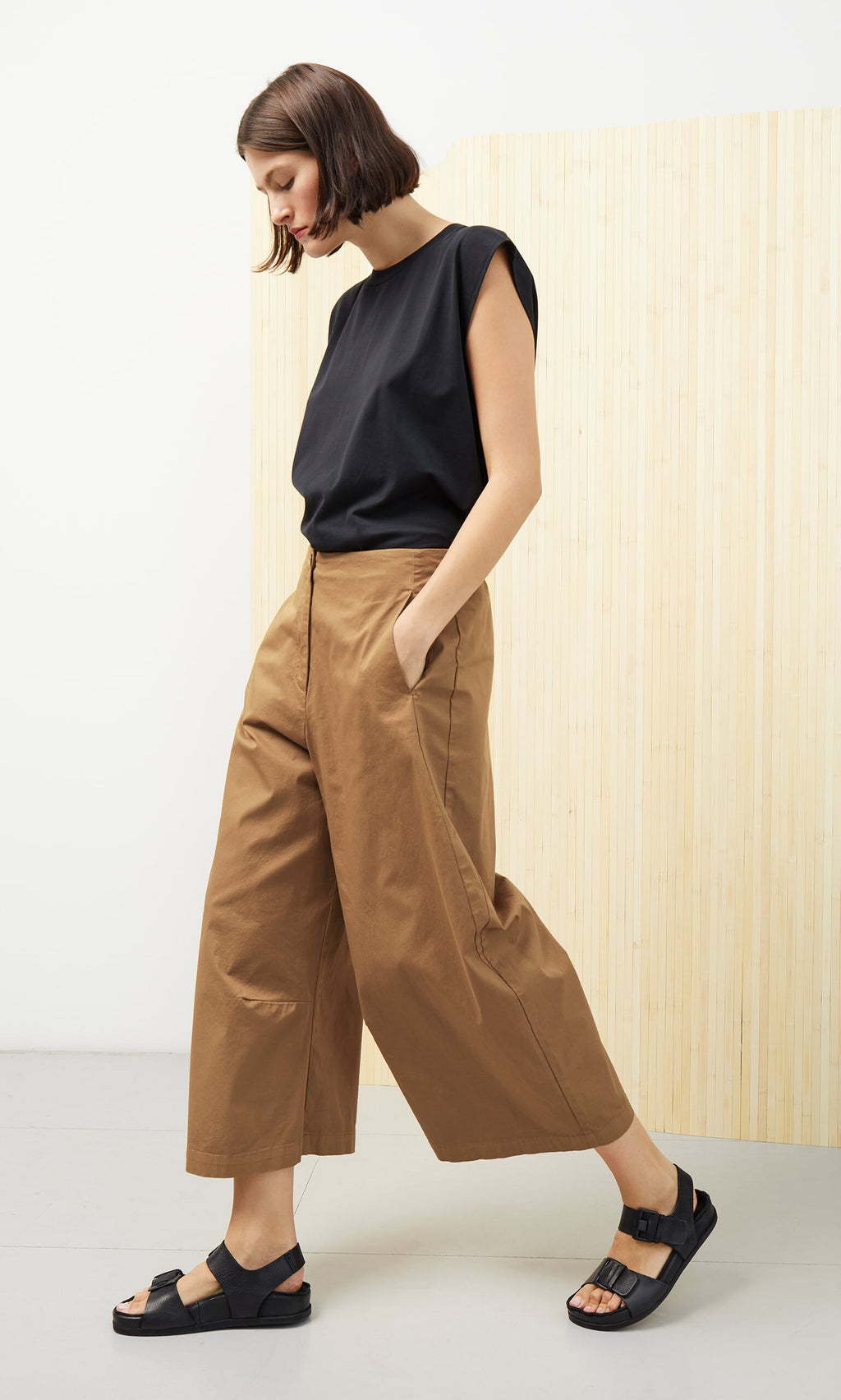 Plümo- Woman wearing black sleeveless top, Karate Pants, and black sandals walks by a light wood panel wall.