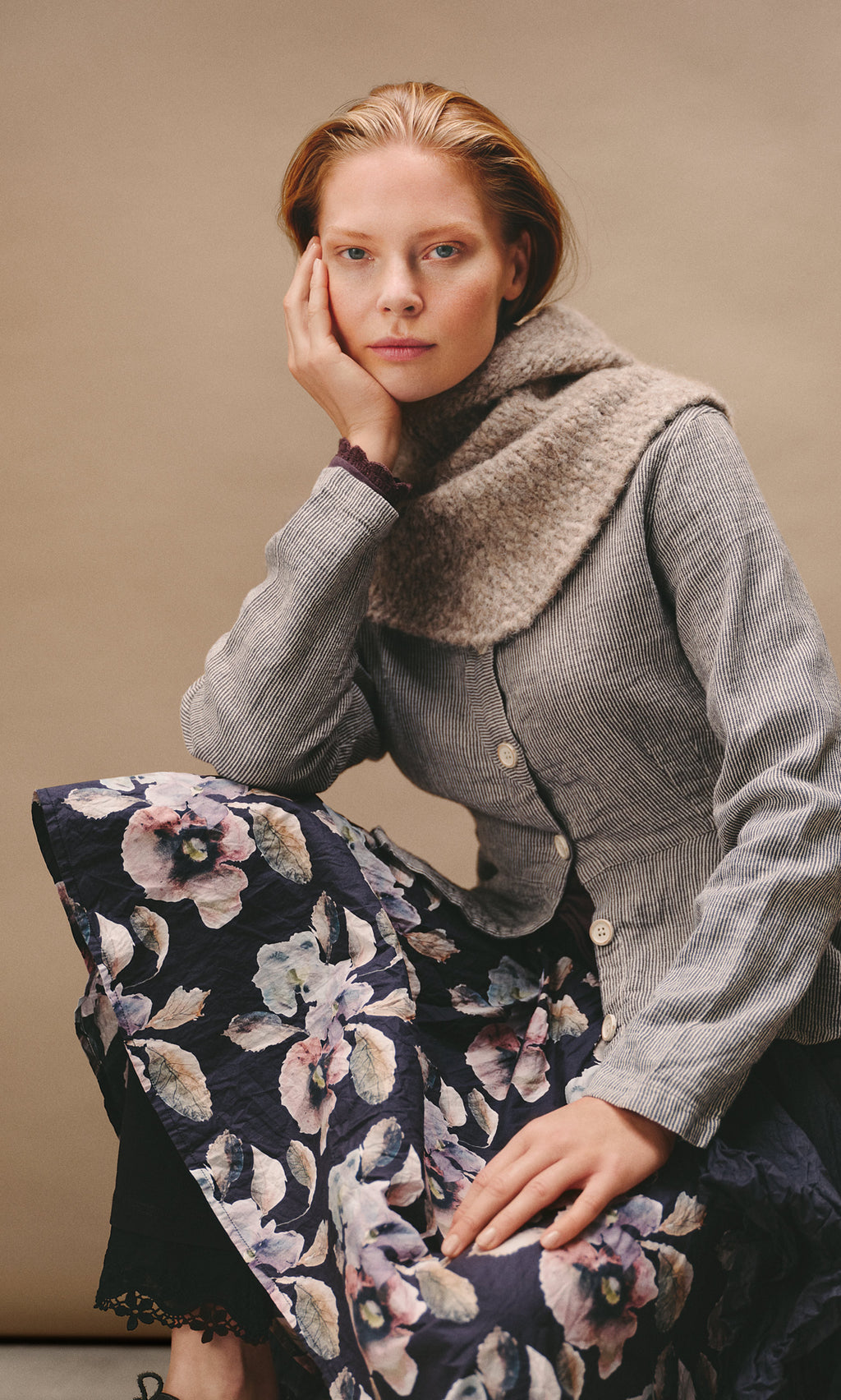 Plümo  A woman wears a dark floral Pugh Flower Skirt, a button-down pinstriped jacket, and a fuzzy cowl neck scarf.
