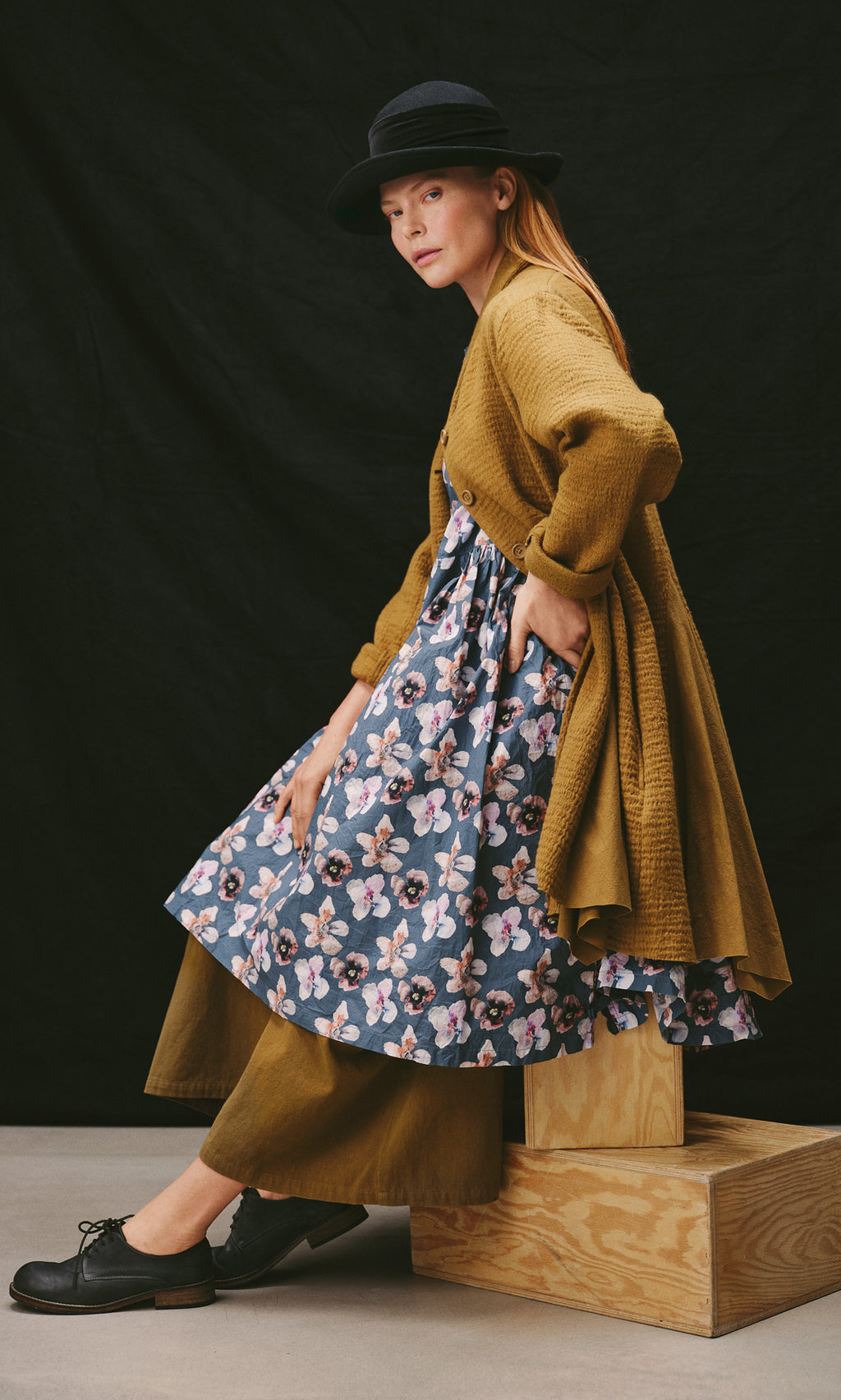 Plümo  A person wearing a Pugh Flower Skirt, olive cardigan, and hat sits on a wooden crate.
