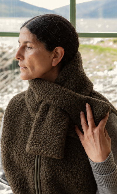 Plümo  Woman with dark hair in the Fern Scarf and a vest, looking left outdoors by a window, hand resting on her scarf.