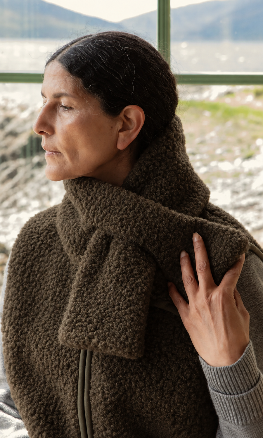 Plümo  Woman with dark hair in the Fern Scarf and a vest, looking left outdoors by a window, hand resting on her scarf.