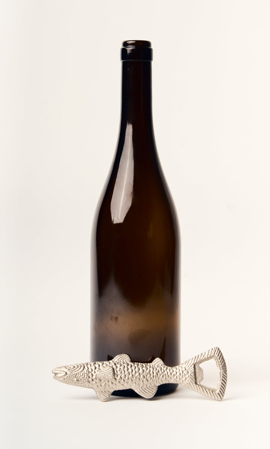Plümo- A Fish Bottle Opener is displayed in front of an upright dark wine bottle on a white background.