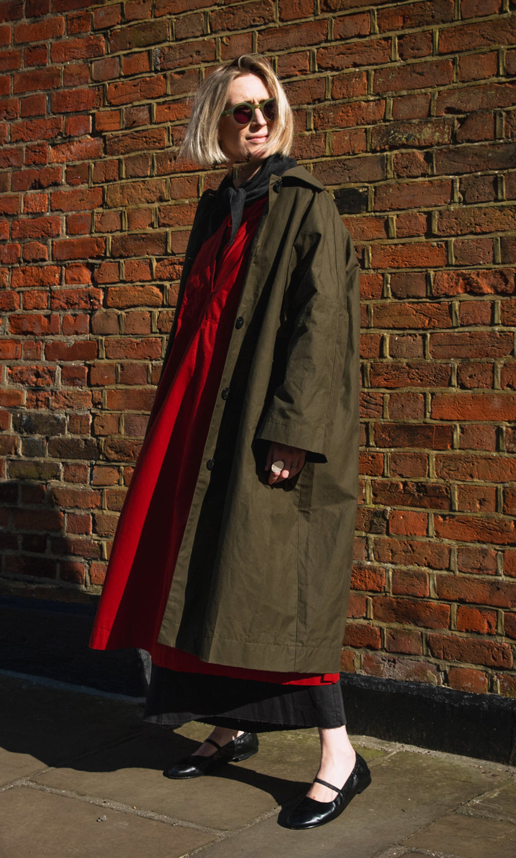 Plümo- Woman in sunglasses, red dress, and Doherty Coat - Olive stands on a sunlit sidewalk by a brick wall.