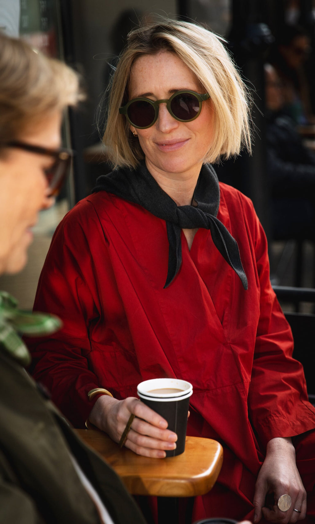 Plümo- A woman in a red coat and Jona Necktie - Graphite smiles with a coffee cup at an outdoor café.