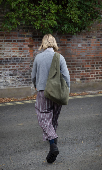 Plümo  A person in a grey sweater and striped trousers carries a large, slouchy olive green Olivia Suede Bag.