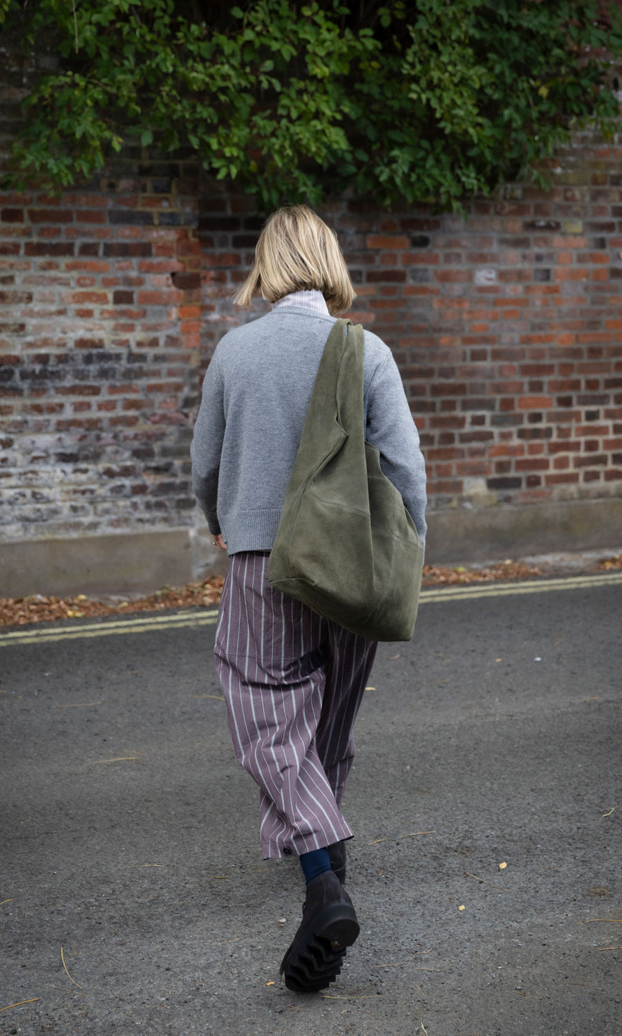 Plümo  A person in a grey sweater and striped trousers carries a large, slouchy olive green Olivia Suede Bag.