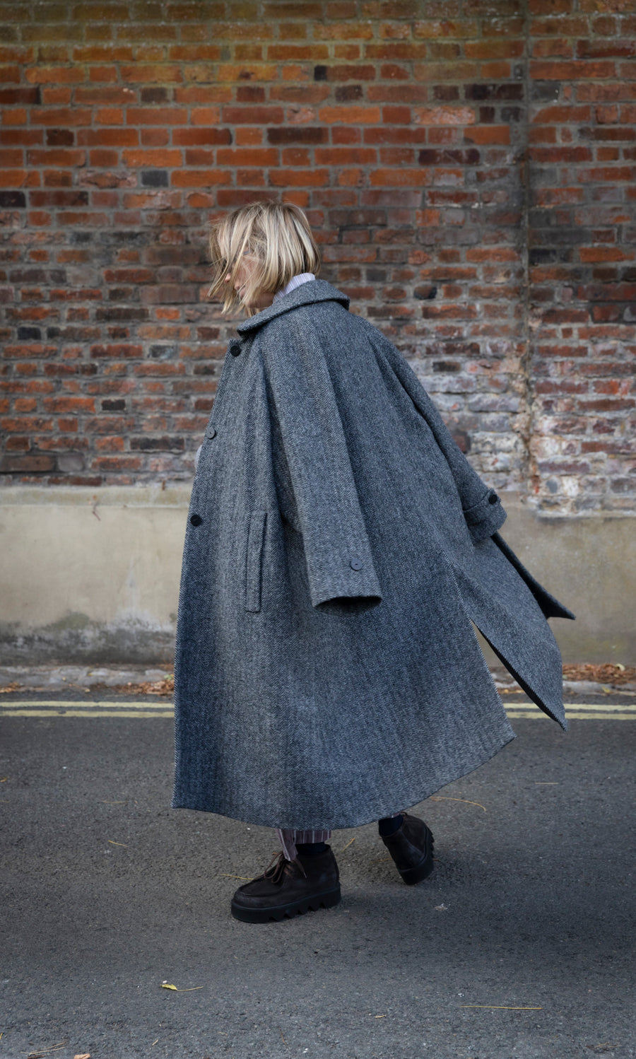 Plümo  A person wearing a grey herringbone Balmacaan Tweed Coat walks past a brick wall.