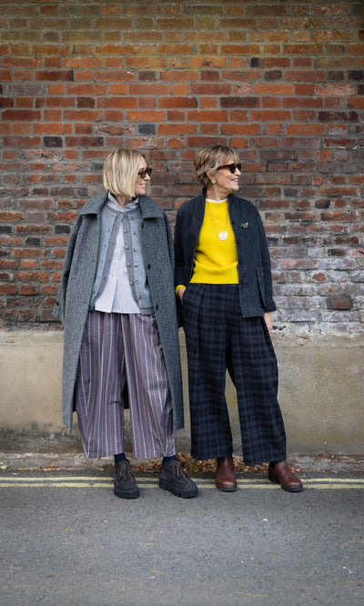 Plümo  Two women in long coats and striped Butoni Pants - Liquorice stand in front of a brick wall.