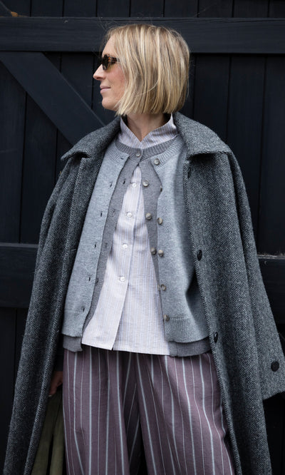 Plümo  A person in a grey herringbone Balmacaan Tweed Coat over a cardigan, striped shirt, and striped wide-leg trousers.