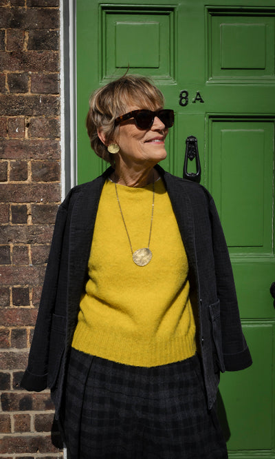 Plümo  A woman wears a Wicklow Crewneck - Golden Hop in mustard yellow, a dark jacket, and a gold circular pendant necklace.