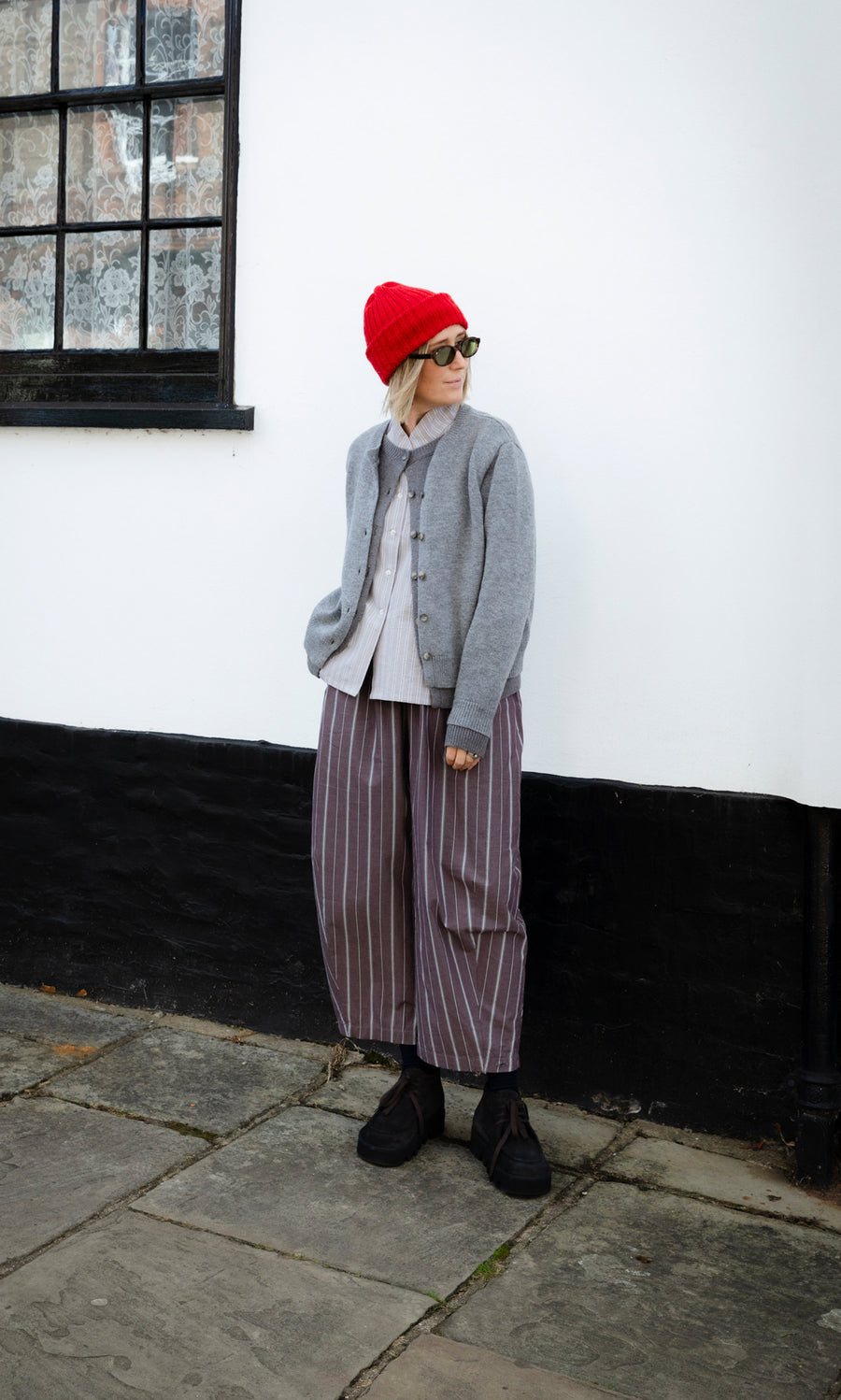 Plümo  Person in a red beanie, grey cardigan, striped button-up shirt, and Butoni Pants - Liquorice.
