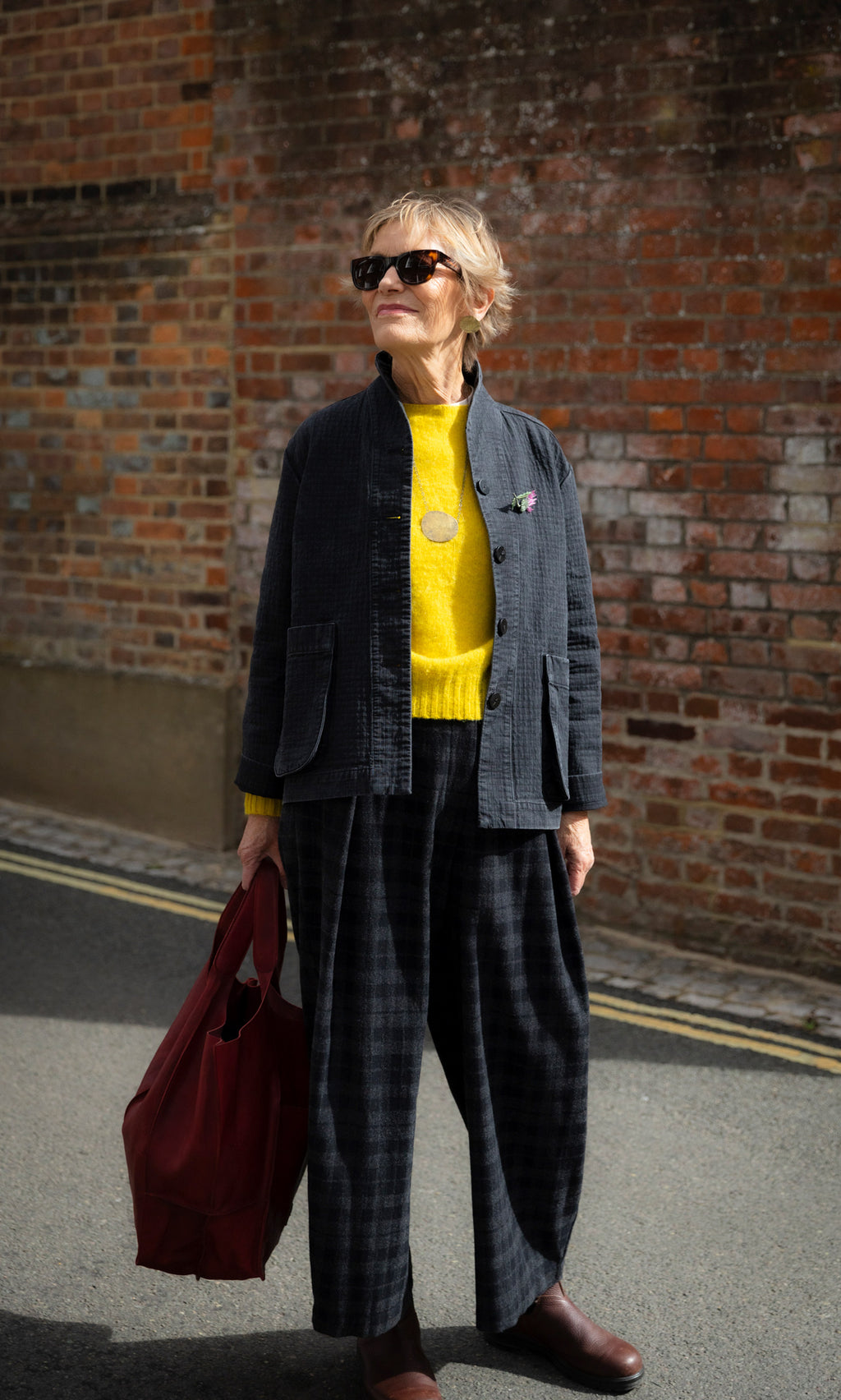 Plümo  A woman in a dark jacket, yellow sweater, and plaid Tartan Flannel Pants stands outdoors holding a large red bag.