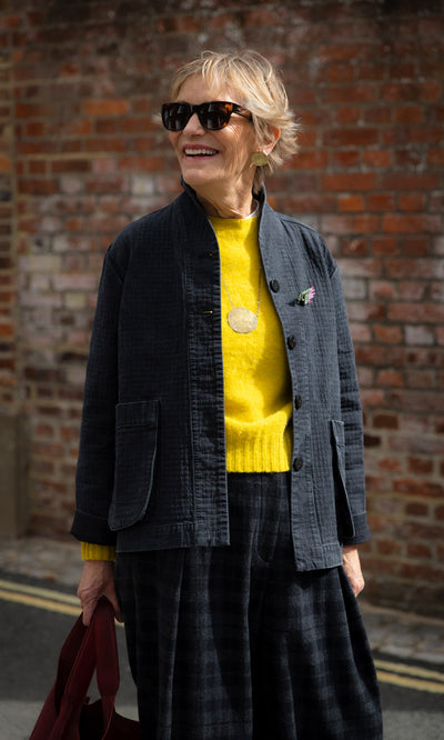 Plümo  A woman in a yellow Wicklow Crewneck - Golden Hop, dark jacket, and plaid trousers standing by a brick wall.
