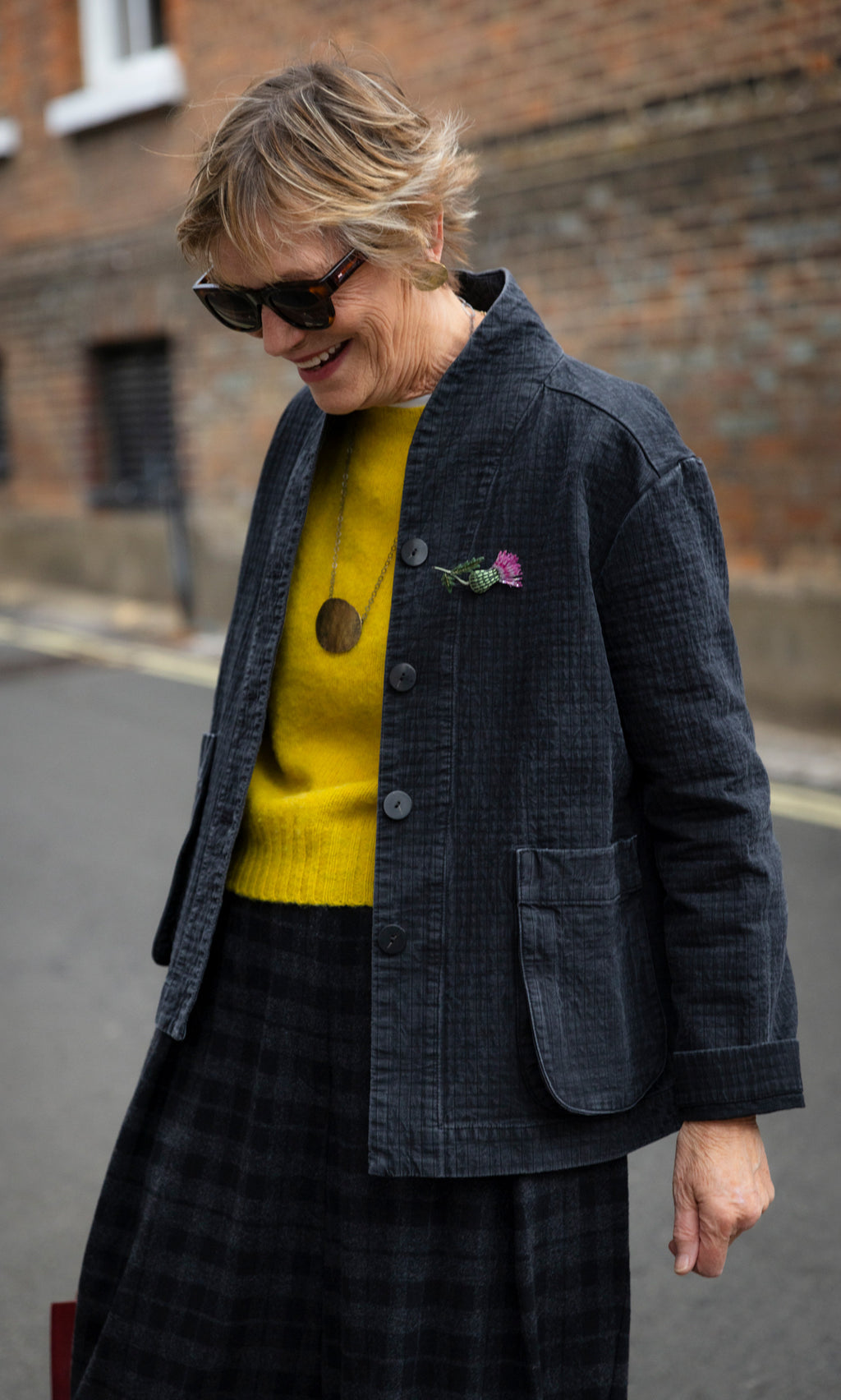 Plümo  Woman wearing a coal Envoy Jacket over a yellow sweater and a plaid skirt.