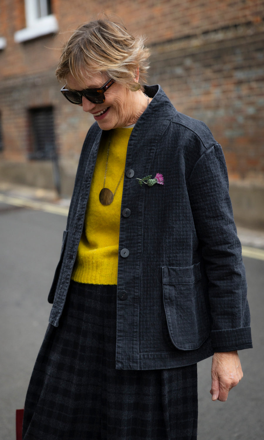 Plümo  Woman wearing a coal Envoy Jacket over a yellow sweater and a plaid skirt.