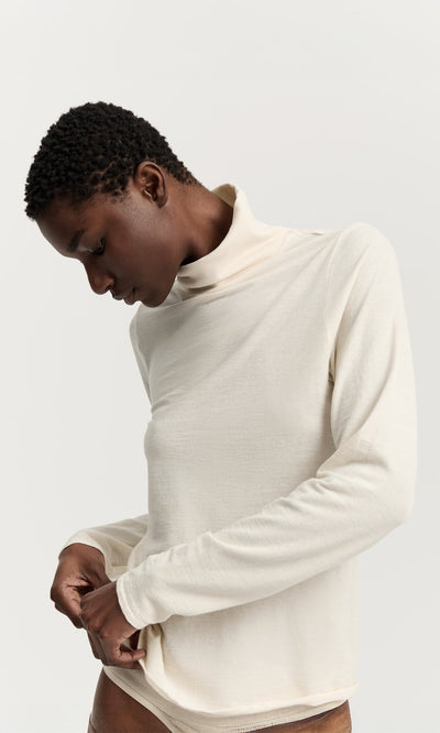 Plümo- Person wearing a Gentle Turtleneck in cream, looking down and adjusting the hem against a plain background.
