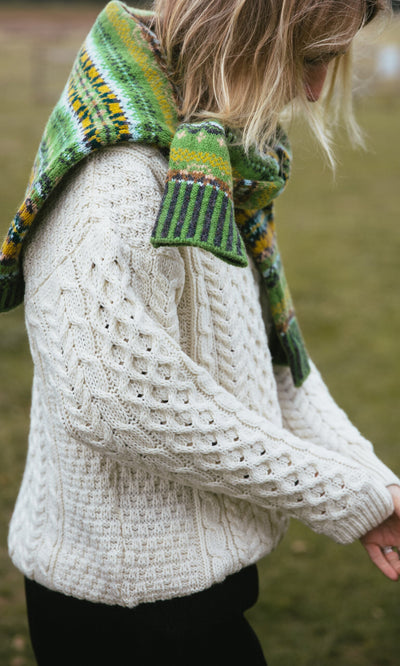 Plümo- Person wearing the Berg Aran Sweater, a natural wool cable knit, with a green patterned scarf outdoors on grass.