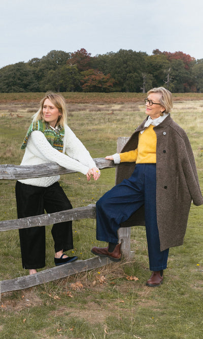 Plümo- Two women in fall outfits, one in the Berg Aran Sweater, lean on a wooden fence in a grassy, tree-lined field.