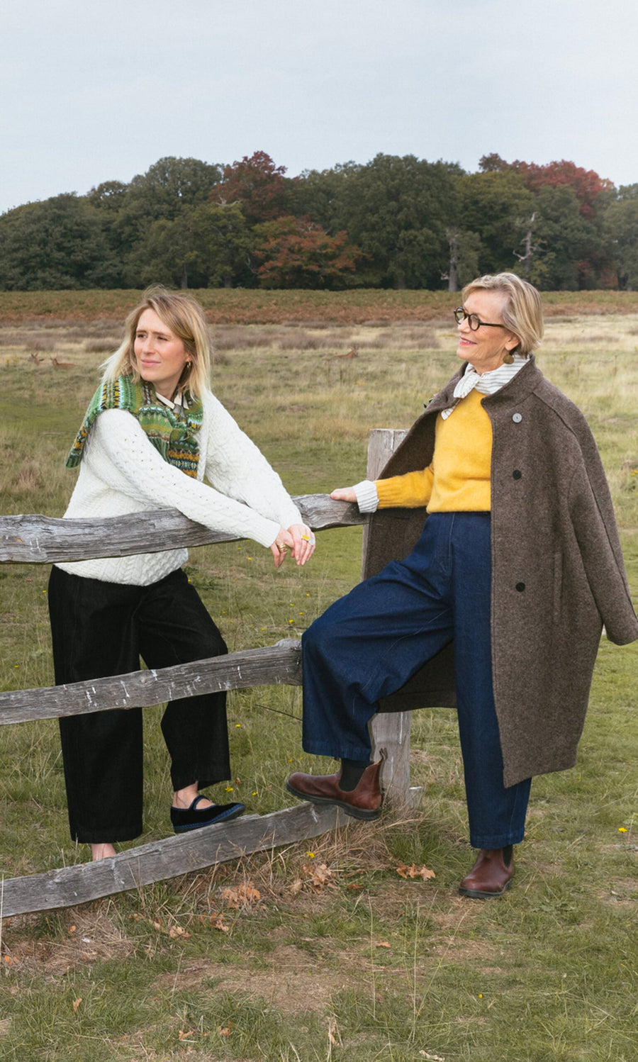 Plümo- Two women in fall outfits, one in the Berg Aran Sweater, lean on a wooden fence in a grassy, tree-lined field.