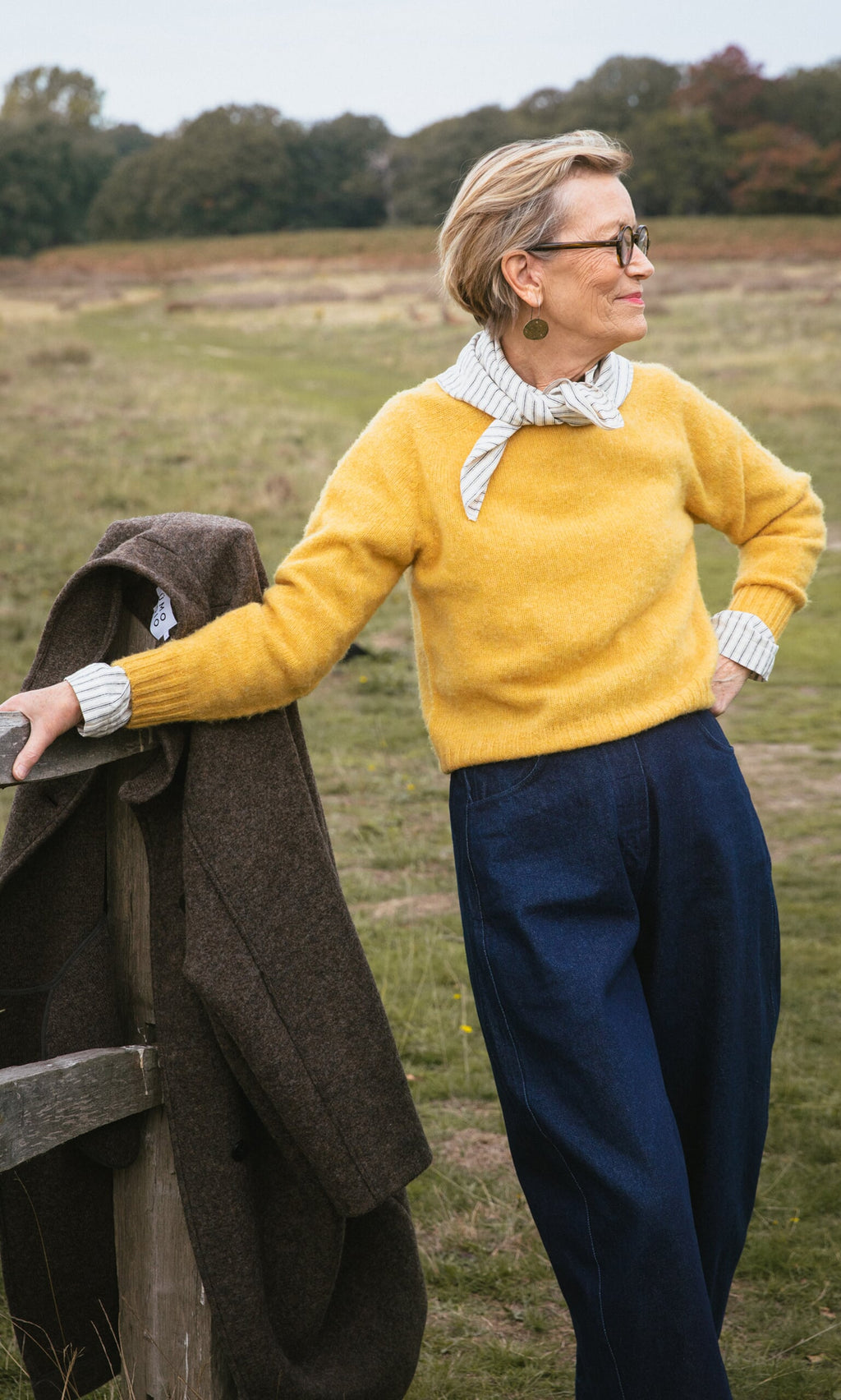 Plümo- Older woman in glasses stands by a wooden fence in a grassy field wearing the Oban Sweater.