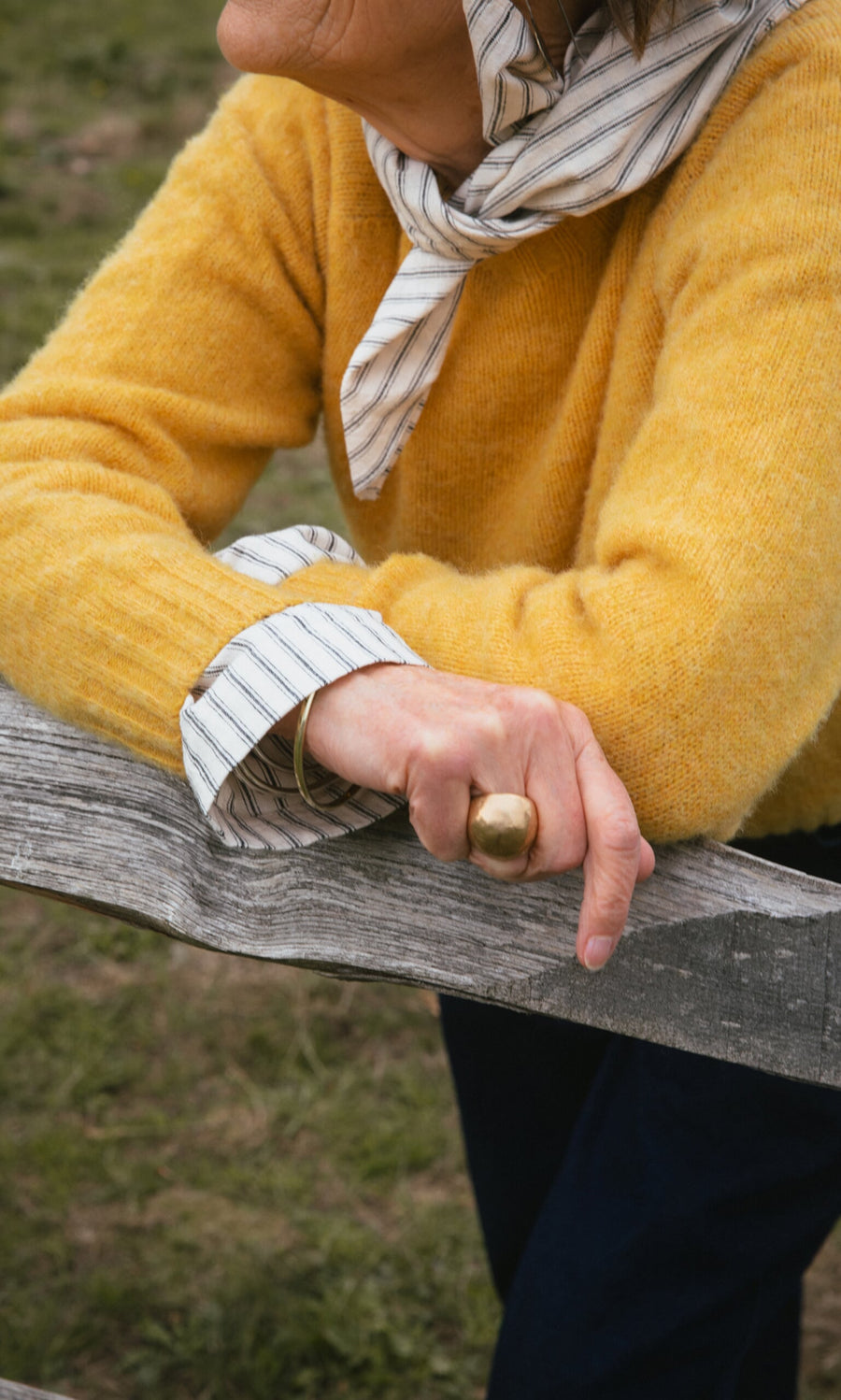 Plümo- A person in a yellow sweater leans on a wooden fence, wearing a striped scarf and the Bolla Ring.