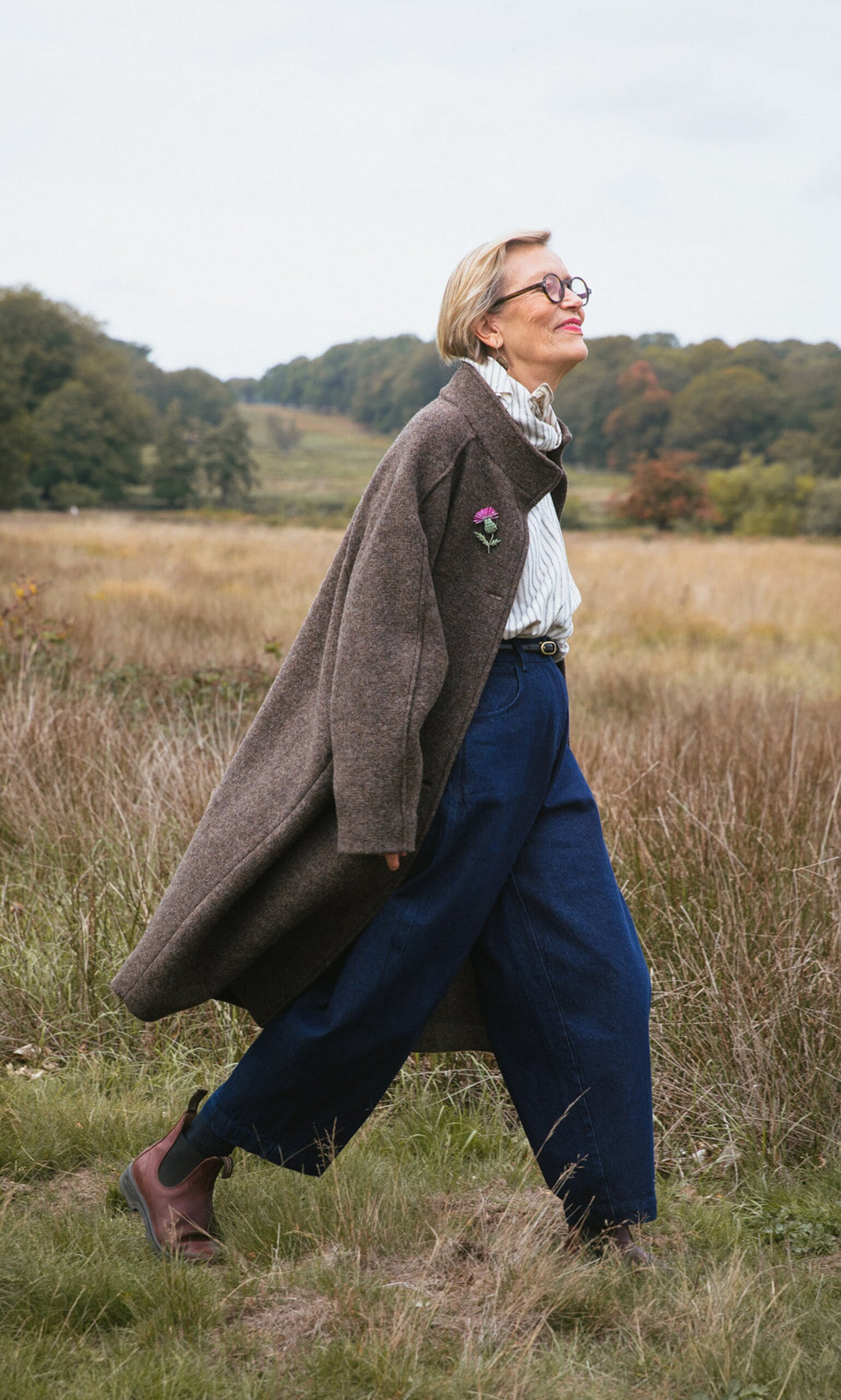 Plümo- Smiling older woman in a Babette Coat, wide-leg jeans, and boots walking through a grassy field.