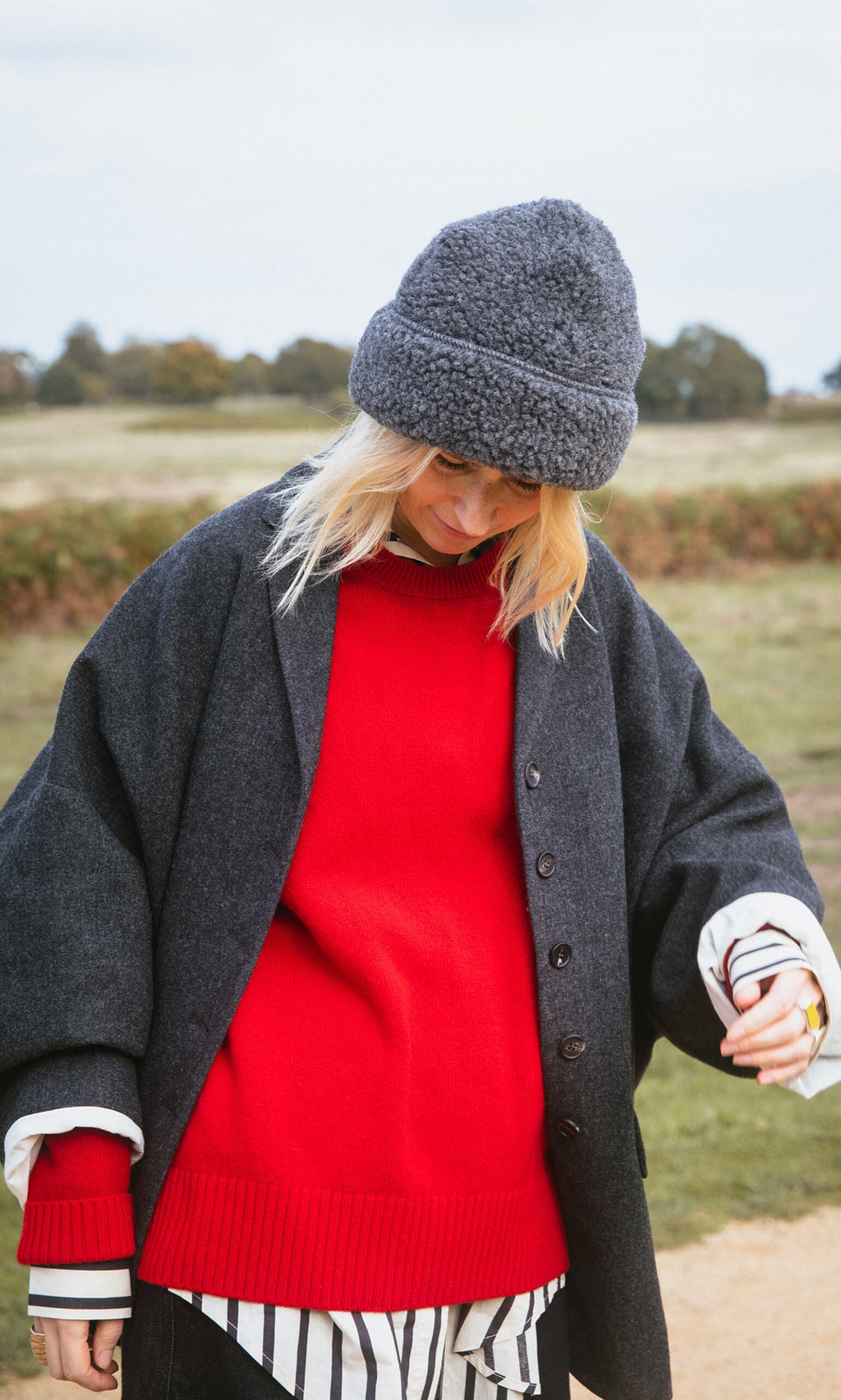 Plümo- Person in a red Bute Sweater, dark coat, and gray hat stands outdoors with a field in the background.