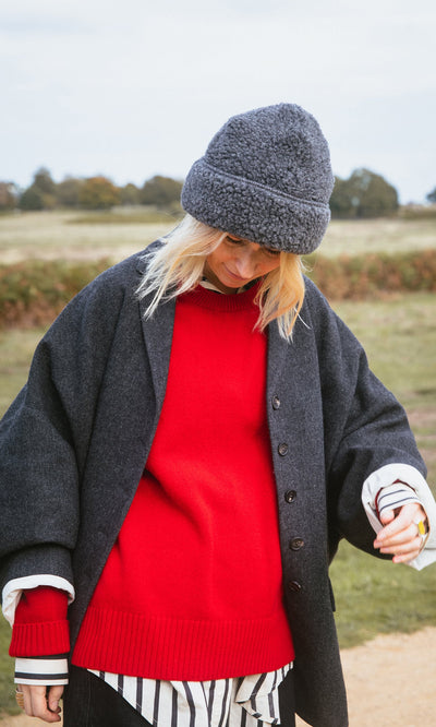 Plümo- Person in a red Bute Sweater, dark coat, and gray hat stands outdoors with a field in the background.
