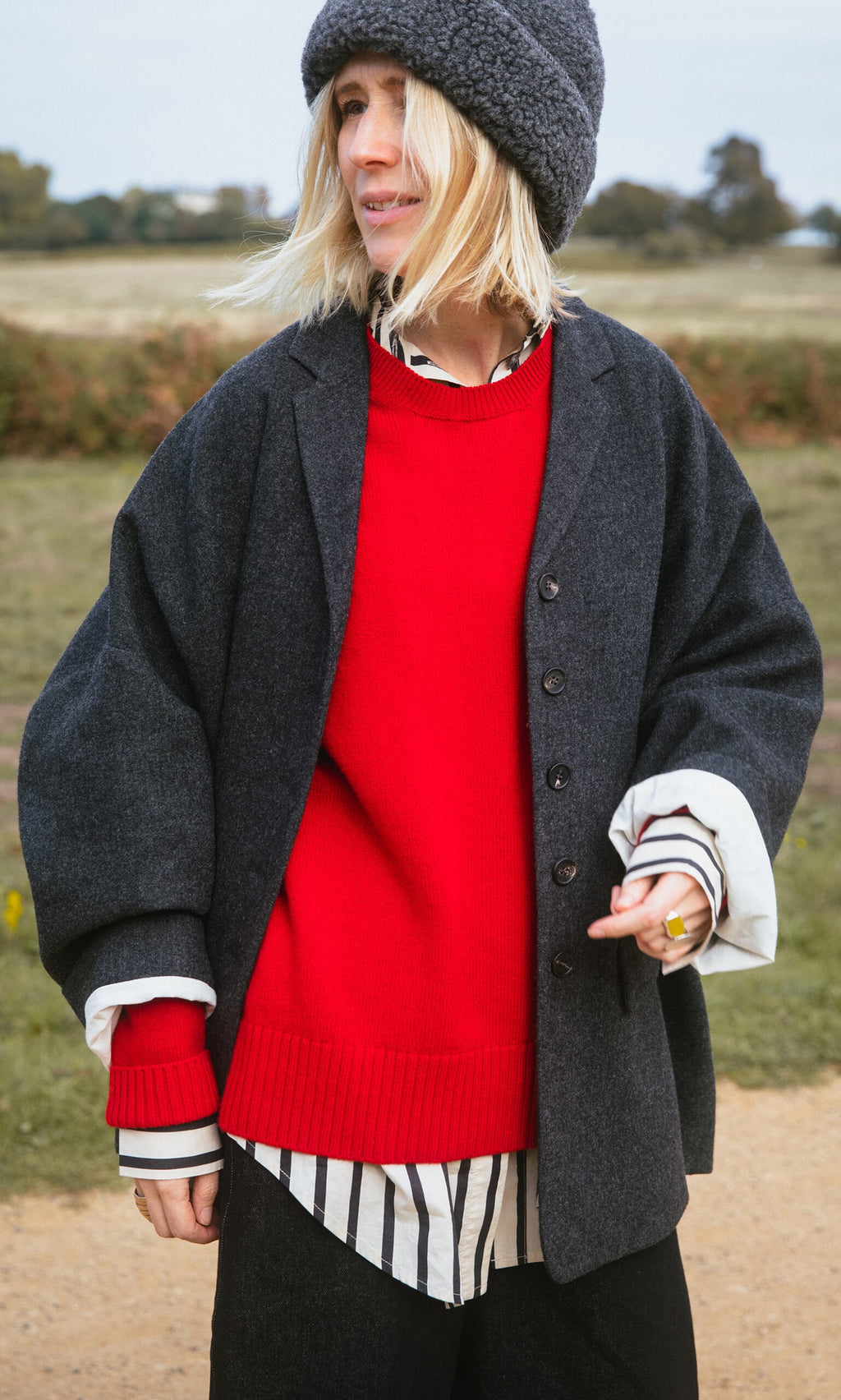 Plümo- Person in a gray coat, Bute Sweater in red, striped shirt, and gray hat standing outdoors on a cloudy day.
