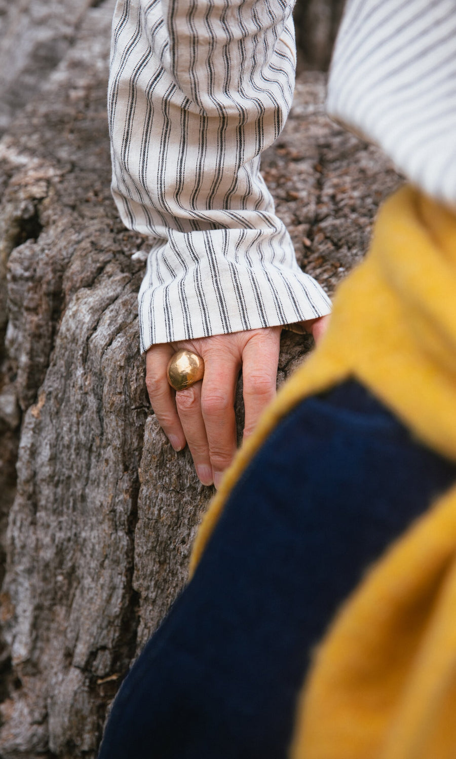 Plümo- Person wearing a striped shirt and Bolla Ring rests hand on tree trunk, yellow and navy fabric in foreground.