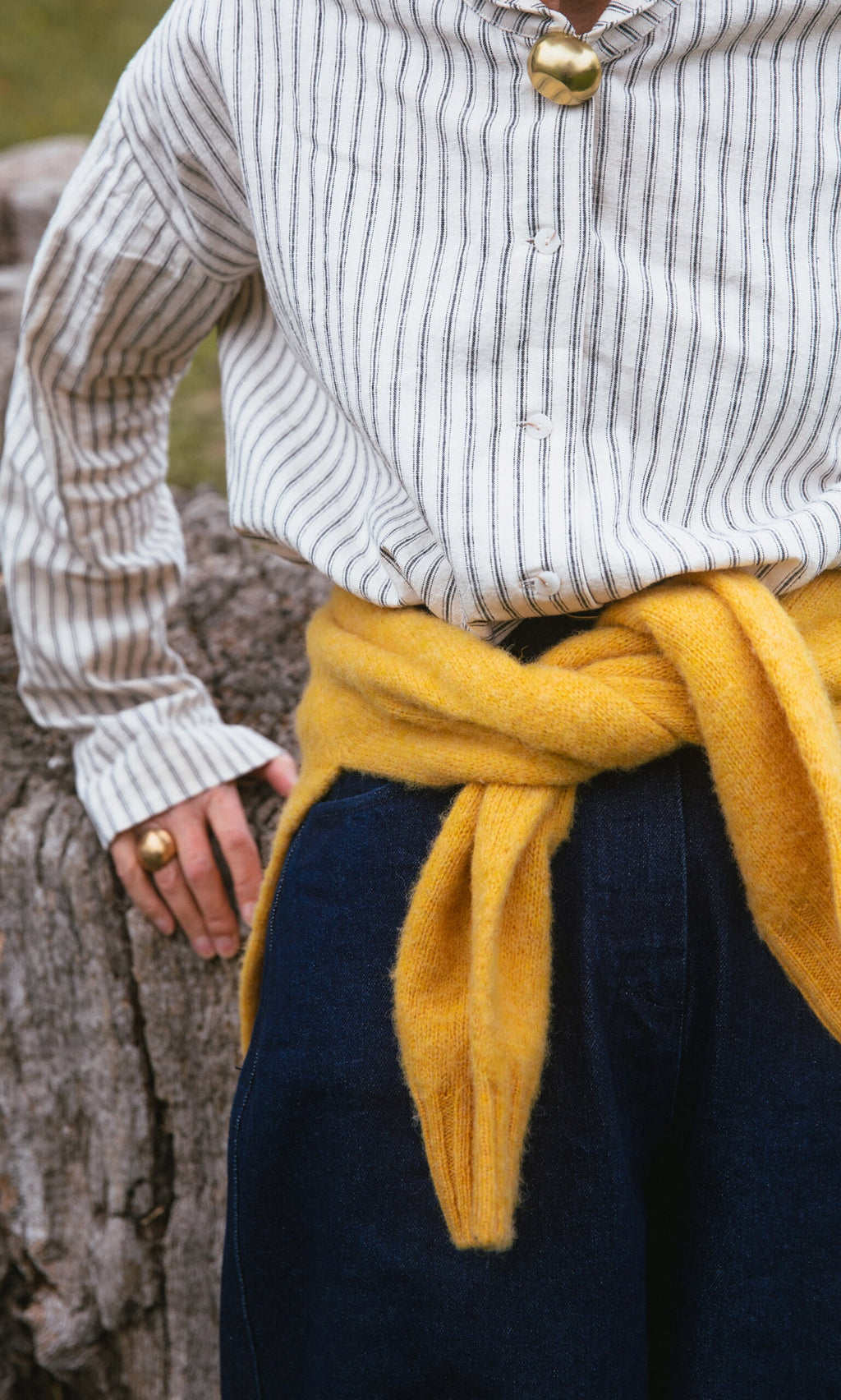 Plümo- Person in striped shirt and yellow sweater with Amara Brooch, standing near a tree trunk.