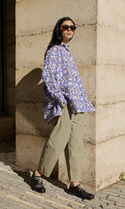 Plümo- Person in sunglasses, floral shirt, and Khaki Poplin Pants stands by a stone wall in sunlight.