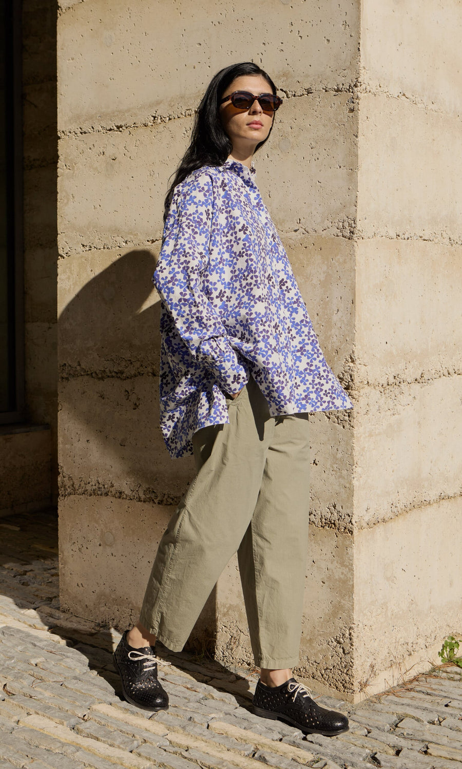 Plümo- Person in sunglasses, floral shirt, and Khaki Poplin Pants stands by a stone wall in sunlight.