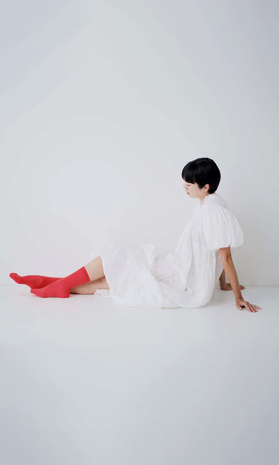 Plümo- Person in a white dress and Japanese Red Socks sits on the floor against a plain white background.