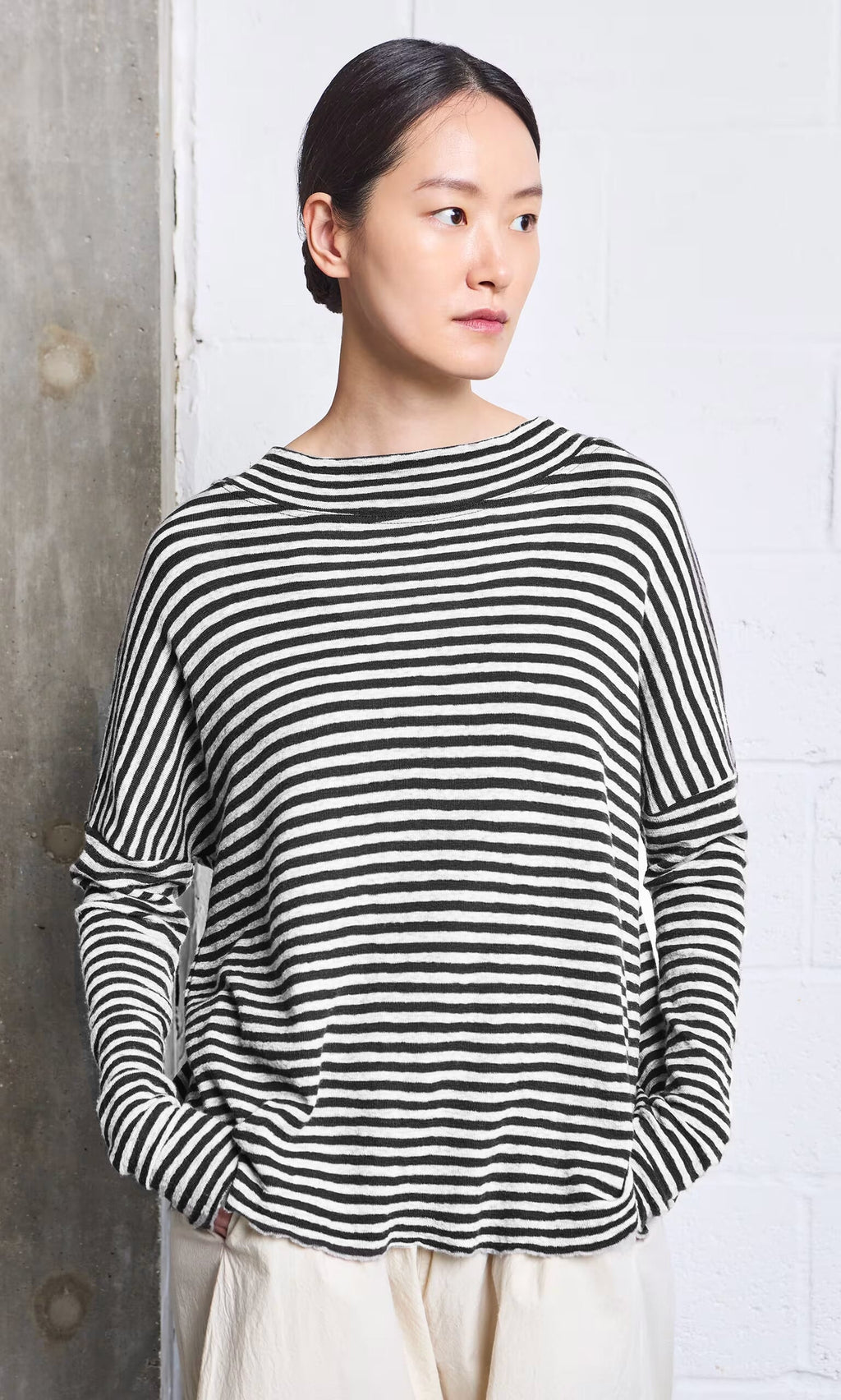 Plümo- Woman wears the Ida Mockneck - Black  White with light pants, standing by a white and concrete wall.