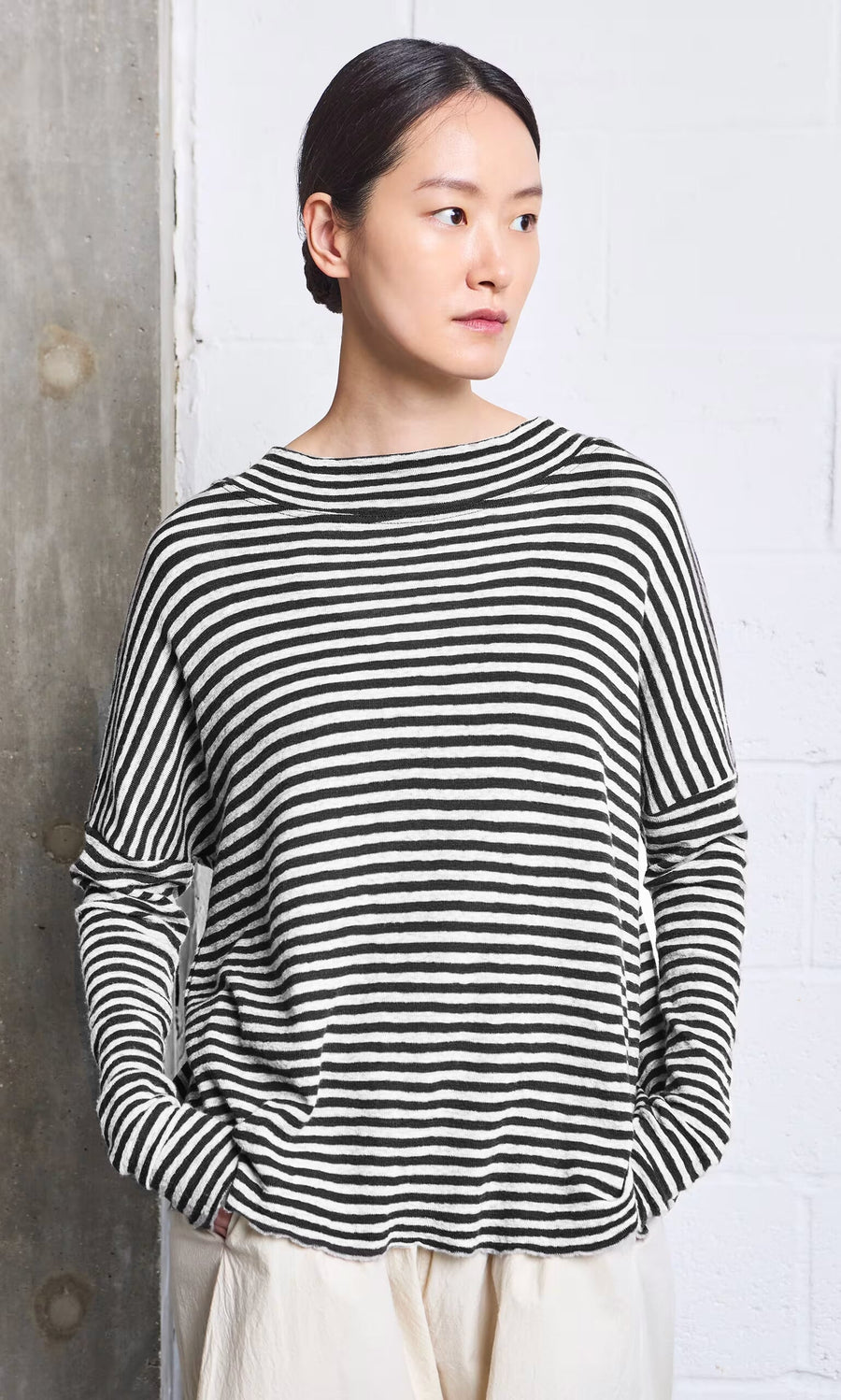 Plümo- Woman wears the Ida Mockneck - Black  White with light pants, standing by a white and concrete wall.