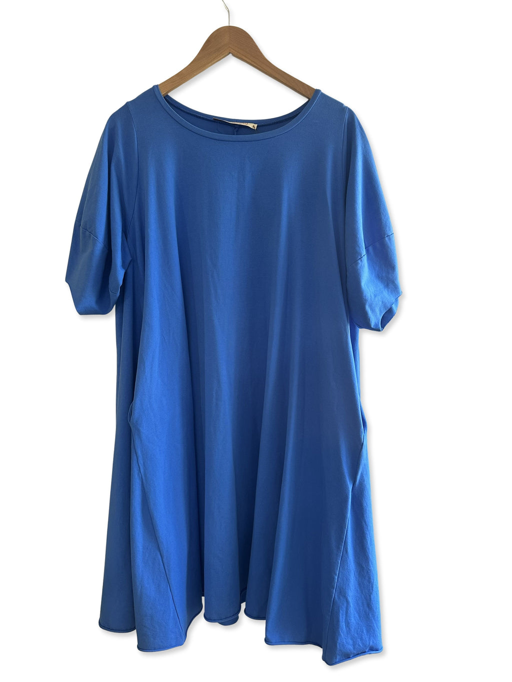 Plümo  The Bevell Dress, a bright blue oversized T-shirt dress with side pockets, hangs on a wooden hanger.