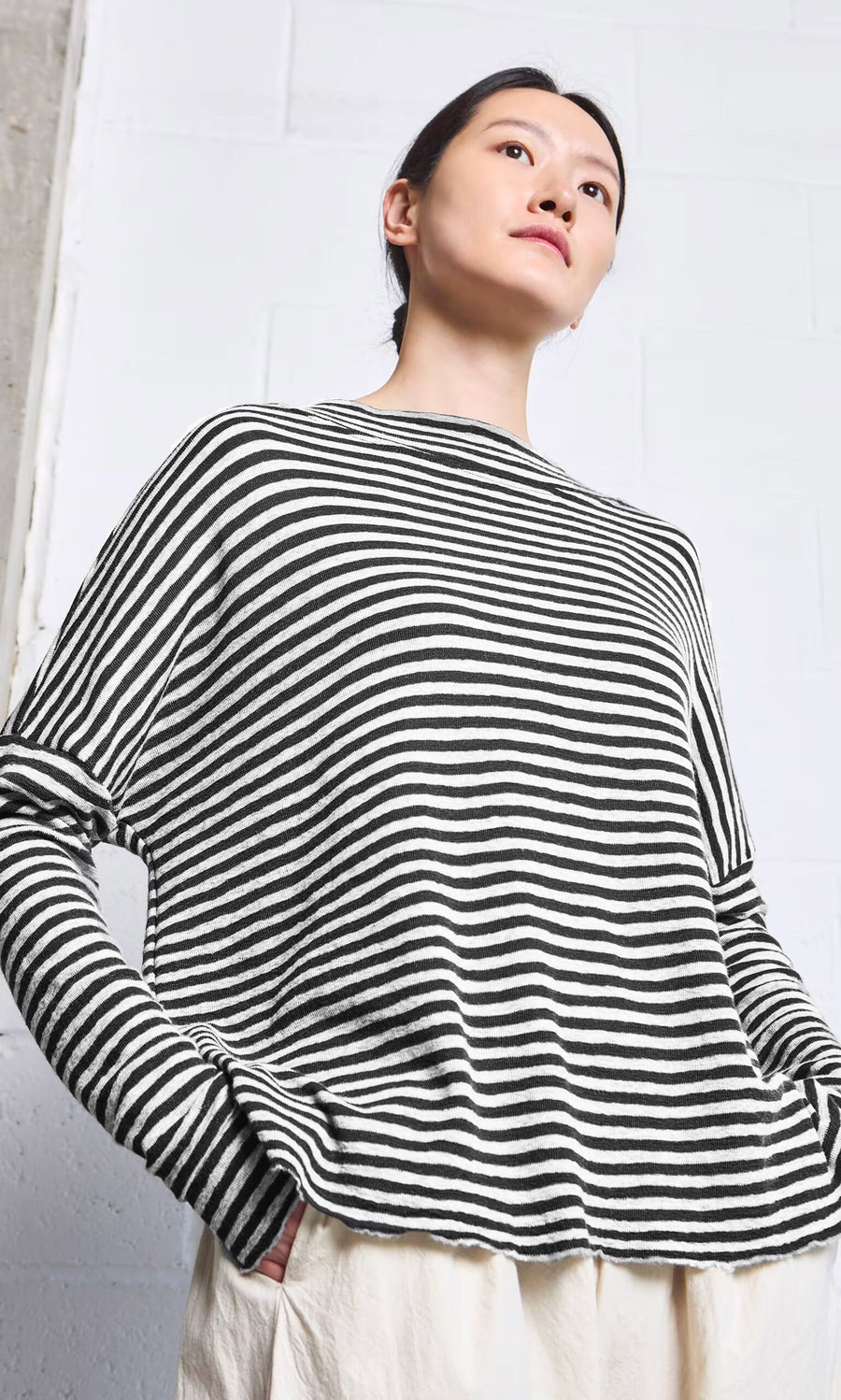 Plümo- Woman wears the Ida Mockneck - Black  White with cream pants, standing with hands in pockets and looking upward.