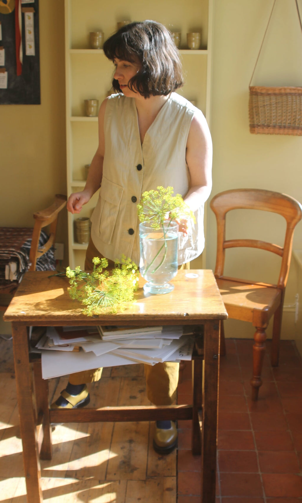 Plümo- A person in a beige vest arranges yellow flowers, inspired by Midweek Recipes by Jess Elliott Dennison, on a table.