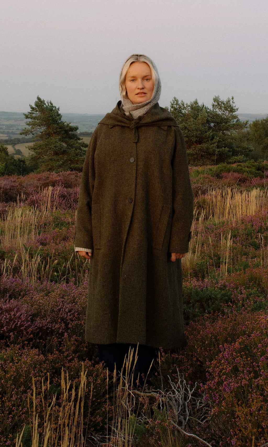 Plümo- Wearing the Cormack Coat - Harris Tweed, a person stands in purple heather fields in the Scottish Outer Hebrides.