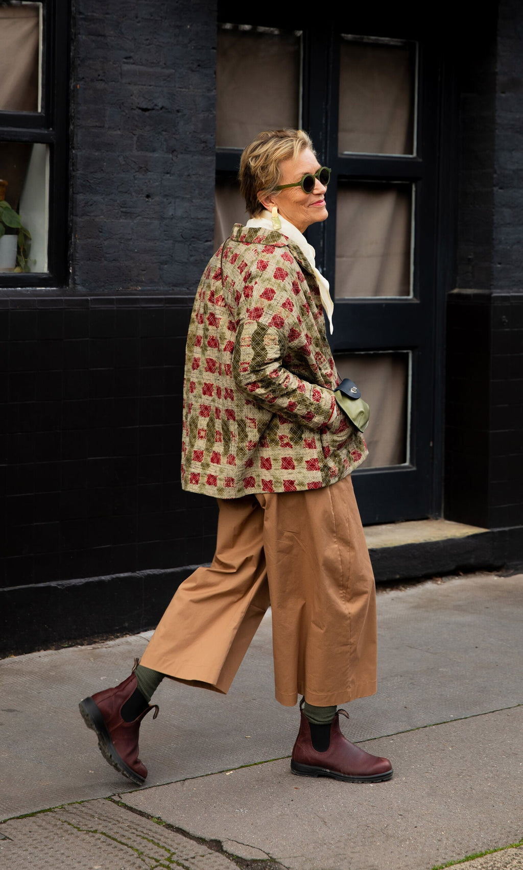Plümo- Person wearing Karate Pants, a patterned jacket, and boots walks on a city sidewalk, looking back over their shoulder.
