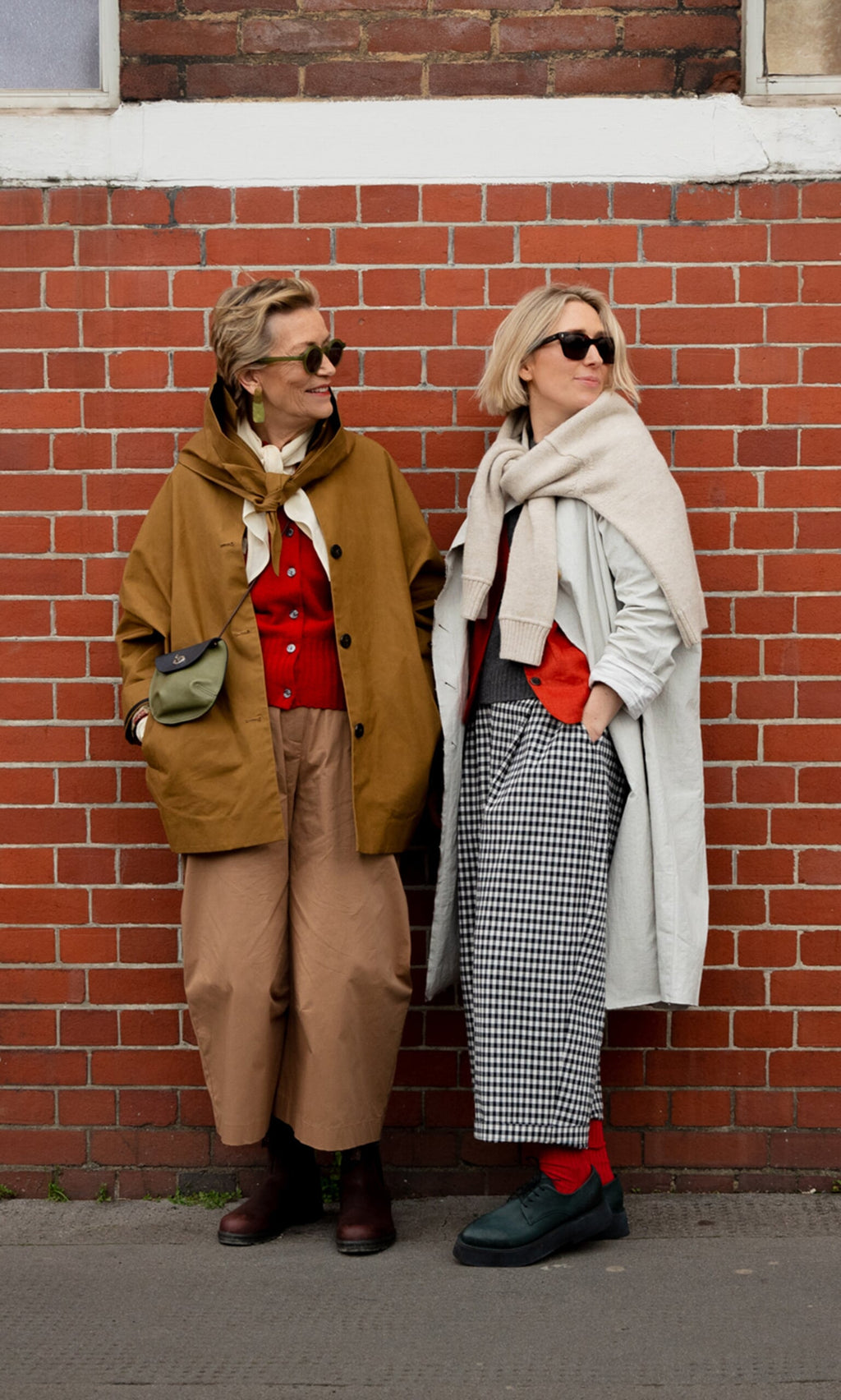Plümo- Two women in stylish layers—one wearing Karate Pants—stand by a red brick wall, both sporting sunglasses.