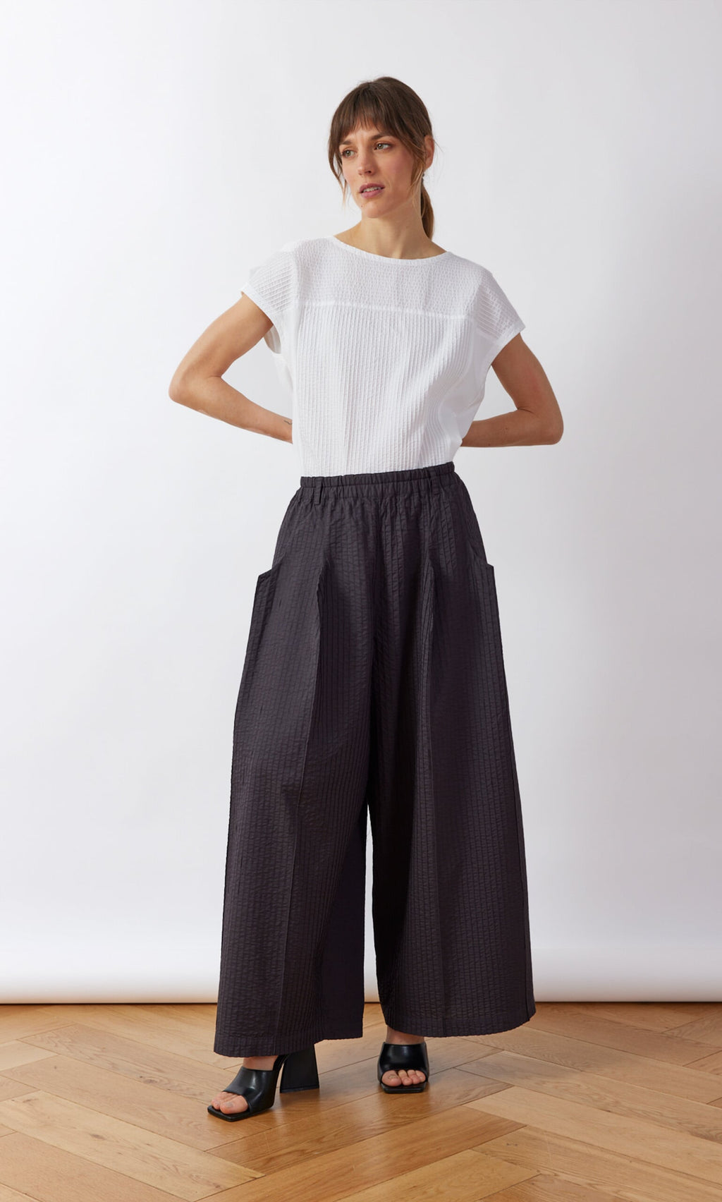 Plümo- Woman in a white short-sleeve top and Kiri Pintuck Pants stands on a wooden floor, hands on hips.