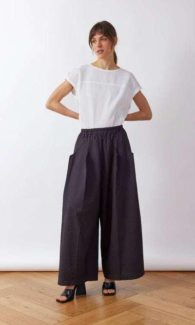 Plümo- Woman in a white short-sleeve top and Kiri Pintuck Pants stands on a wooden floor, hands on hips.