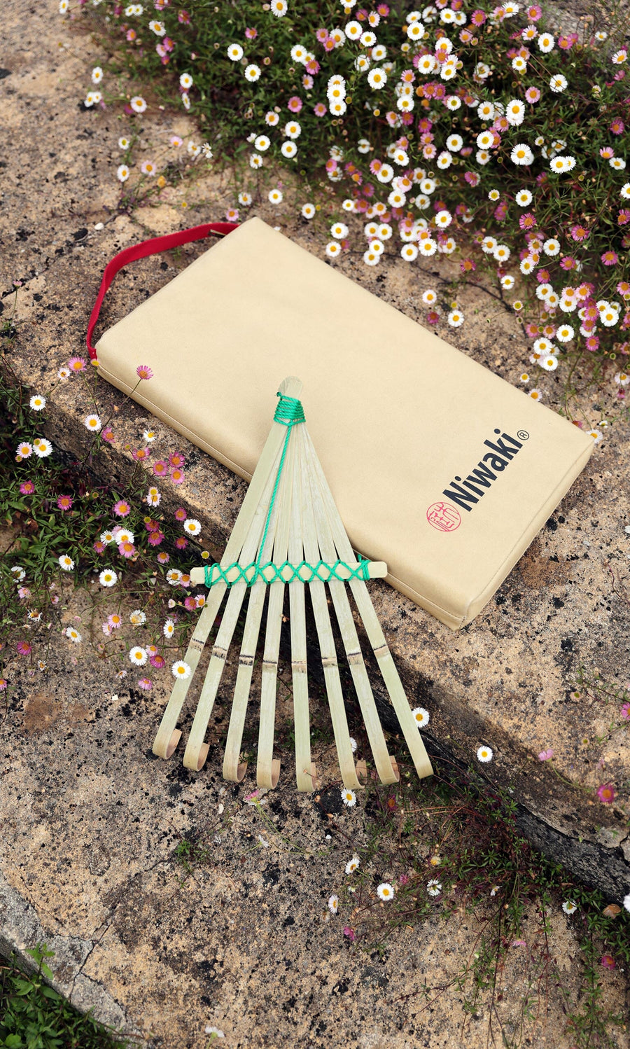 Plümo- A Niwaki Kneeler with a bamboo hand rake and beige carrying case rests among white and pink flowers.
