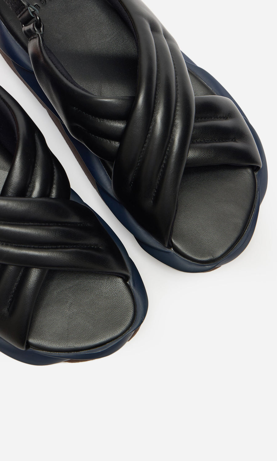 Plümo- Close-up of Korra Sandals—black padded leather with crisscross straps and thick soles on a white background.