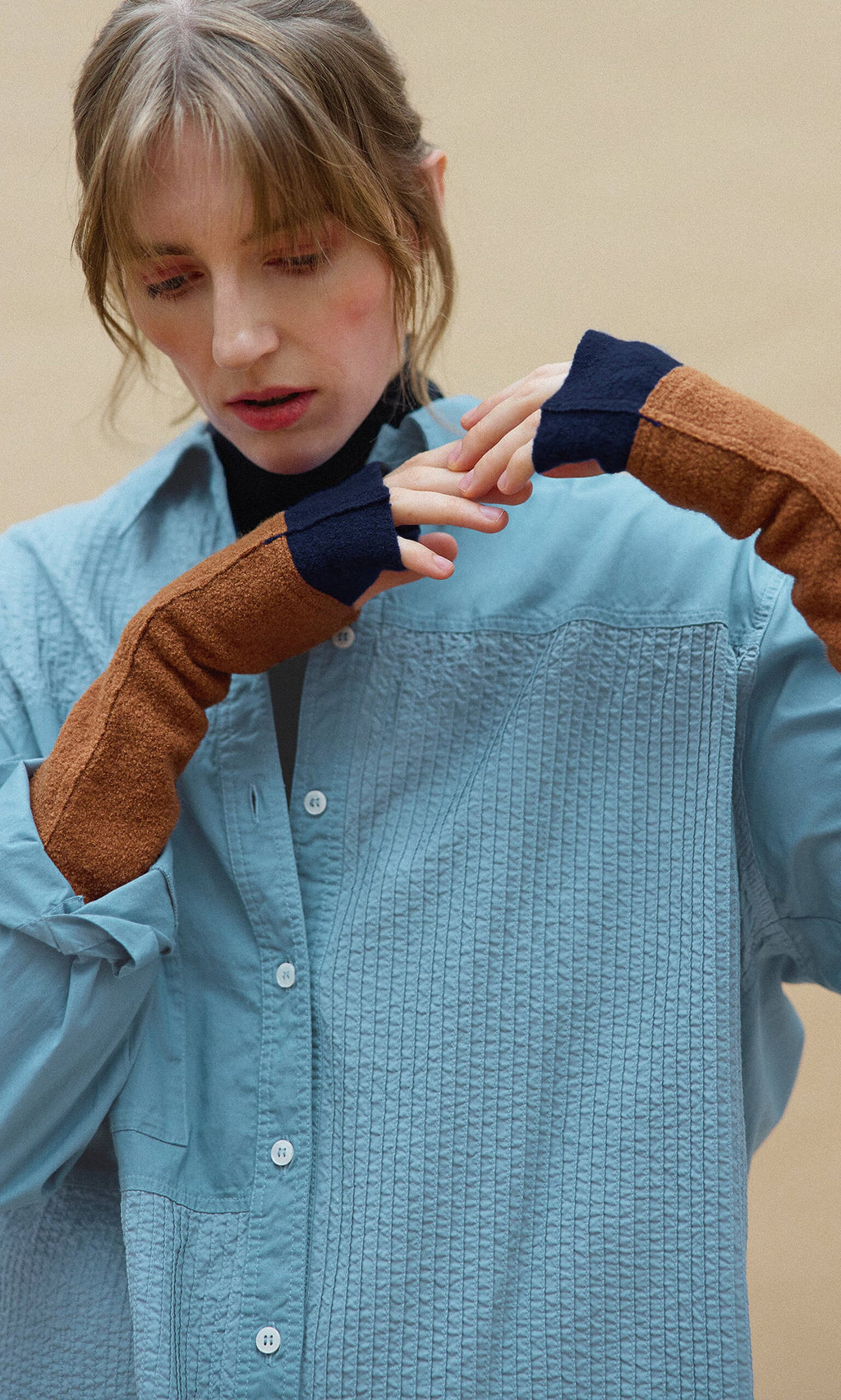 Plümo- Woman in a blue Leung Shirt by Yacco Maricard and long brown gloves looks down, gently touching her hands.