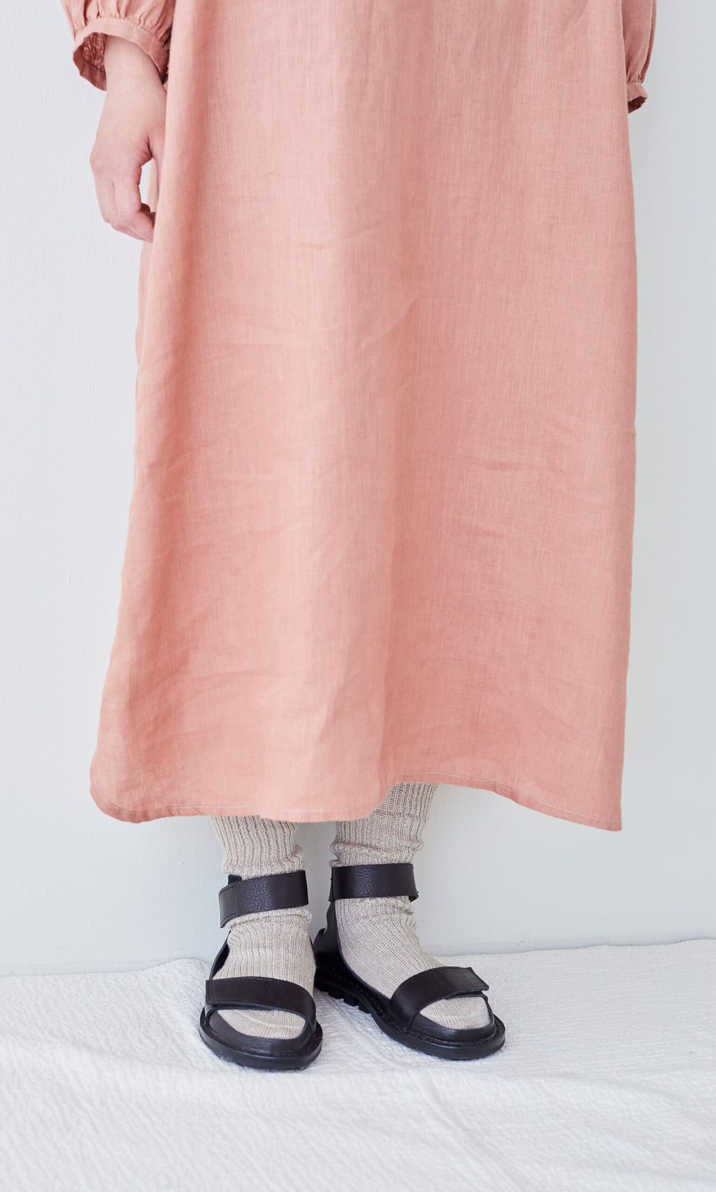 Plümo- Person wearing Linen Rib Socks with a long pink dress and black sandals, standing on a white surface by a plain wall.
