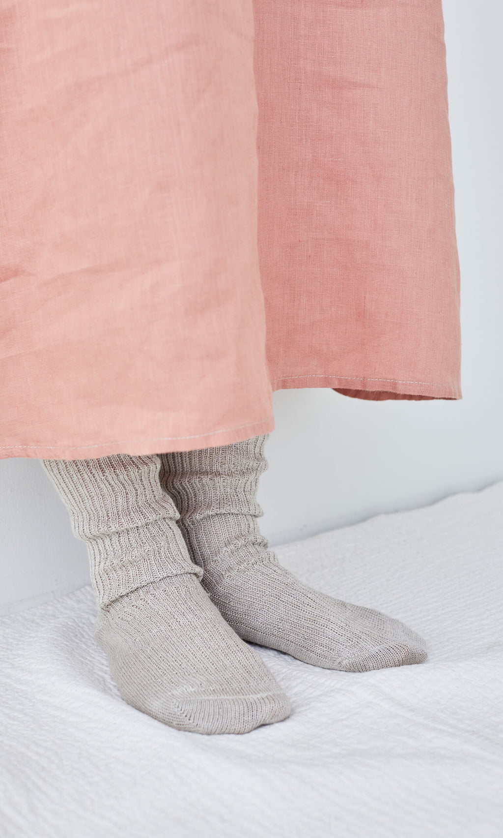 Plümo- Person wearing Linen Rib Socks and a long pink skirt, standing on a white textured surface against a white wall.